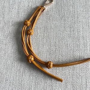 May include: Mustard yellow leather strap with three decorative knots and a gold-colored clasp. The strap is designed for attaching to a bag or other accessory. The knots add a unique design element.