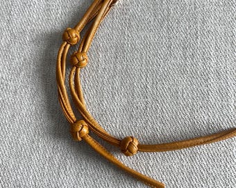 Könnte beinhalten: Senfgelber Lederriemen mit drei dekorativen Knoten und einem goldfarbenen Verschluss. Der Riemen ist zum Befestigen an einer Tasche oder einem anderen Accessoire gedacht. Das Leder hat eine glatte Textur.