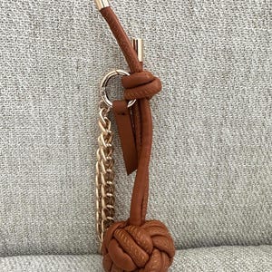 K&ouml;nnte beinhalten: Ein brauner Leder-Schl&uuml;sselanh&auml;nger mit goldfarbener Kette und Akzenten. Der Schl&uuml;sselanh&auml;nger hat unten ein geknotetes Design und eine Schlaufe f&uuml;r Schl&uuml;ssel. Die Oberseite hat eine goldfarbene Metallkappe.
