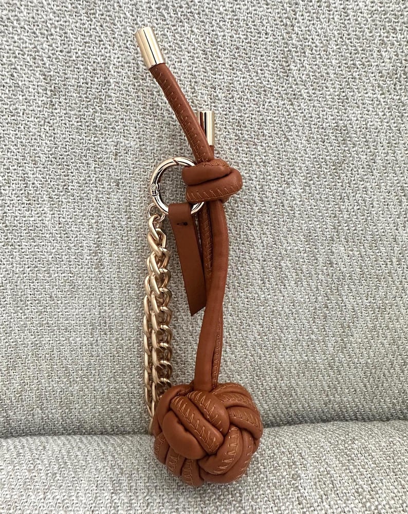 K&ouml;nnte beinhalten: Ein brauner Leder-Schl&uuml;sselanh&auml;nger mit goldfarbener Kette und Akzenten. Der Schl&uuml;sselanh&auml;nger hat unten ein geknotetes Design und eine Schlaufe f&uuml;r Schl&uuml;ssel. Die Oberseite hat eine goldfarbene Metallkappe.