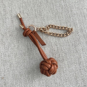K&ouml;nnte beinhalten: Ein brauner Leder-Schl&uuml;sselanh&auml;nger mit dekorativem Knotendesign und goldfarbener Kette. Der Schl&uuml;sselanh&auml;nger hat ein kleines Lederetikett und goldfarbene Metallakzente. Der Knoten ist aufwendig gewebt und verleiht dem Accessoire eine einzigartige Textur.
