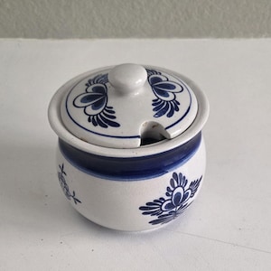 Puede incluir: Un azucarero de cerámica blanca con tapa, con diseños florales azules. El cuenco tiene una banda azul alrededor del centro y una pequeña abertura para una cuchara. La tapa tiene un pomo para facilitar su manejo.