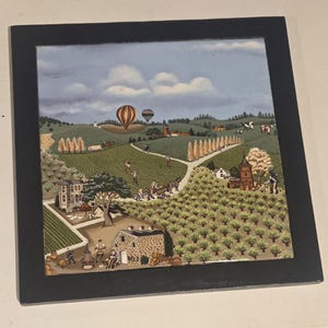 May include: A framed landscape painting depicting a rural scene with rolling green hills, fields, and a winding road. Two hot air balloons float in the sky. The artwork features a black frame and a detailed, colorful illustration.