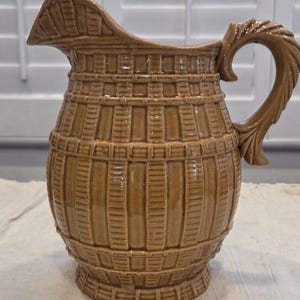 May include: A tan ceramic pitcher with a basket weave design. The pitcher has a curved handle and a spout for pouring. The pitcher is a light brown color and has a glossy finish. The pitcher is a decorative item.