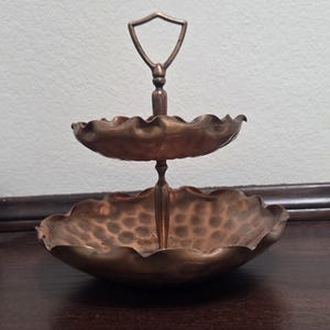 Op de afbeelding: Een tweelaags serveerschaal in koperkleur met een decoratief handvat. De schalen hebben een geschulpte rand en een gehamerde textuur. De handgreep heeft de vorm van een schild. De schaal is ontworpen om kleine items te bevatten.