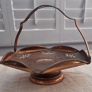 Op de afbeelding: Een koperkleurige decoratieve mand met een handvat. De mand heeft een geschulpte rand en een ronde basis. Witte bloemmotieven zijn aan de binnenkant van de mand geschilderd.