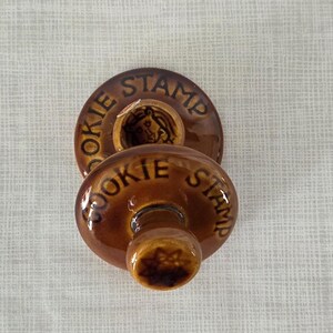 May include: Two round, brown glazed ceramic cookie stamps with a glossy finish. Each stamp features the text COOKIE STAMP embossed in dark brown lettering along the top edge. One stamp shows a star-shaped design on its handle base.