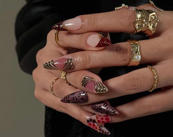 May include: Close-up of a hand with long, pointed, and elaborately decorated nails. The nails feature a variety of designs, including pink, burgundy, and gold accents. Several gold rings adorn the fingers, adding to the overall glamorous aesthetic.