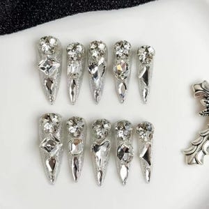 May include: A set of ten silver glitter press-on nails, each adorned with an array of sparkling clear rhinestones in various shapes. The nail tips are pointed, creating a glamorous and eye-catching design. The nails are arranged on a white surface.