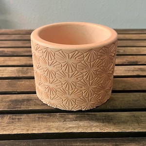 May include: A cylindrical peach-colored planter featuring a repeating geometric pattern of embossed sunburst or star-like shapes across its exterior surface. The vessel has a smooth, thick rim and a flat base, showcasing a consistent matte finish.
