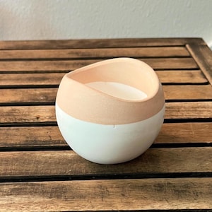 May include: A round, two-toned candle holder with a white base and a peach-colored top. The candle holder is filled with white wax. The object is sitting on a wooden surface.