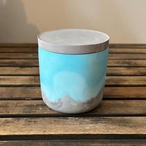 May include: A cylindrical container with a light blue and gray painted design. The container has a matching gray lid. The design on the container resembles a landscape with a blue sky and gray mountains.