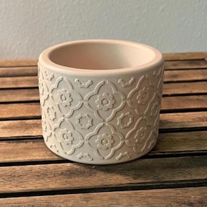 May include: A beige ceramic planter with a repeating floral pattern. The cylindrical pot has a wide opening and sits on a wooden surface. The embossed design adds texture to the neutral-toned pot, suitable for indoor or outdoor use.