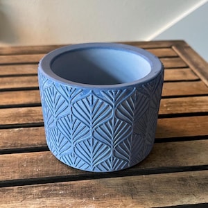 May include: A blue cylindrical planter with a textured leaf pattern. The planter is made of a solid material and sits on a wooden surface. The interior of the planter is also blue.