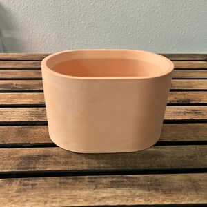 May include: A smooth, oval-shaped planter with a matte finish in a light peach or terracotta color. The vessel features a wide, open top and solid, continuous walls, presenting a clean, uniform appearance without any decorative patterns or textures.