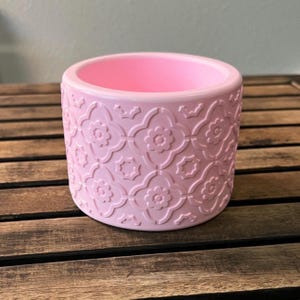 May include: A pink ceramic planter with a floral pattern. The cylindrical pot has a smooth, glossy finish and a wide opening. The embossed design features repeating flower and star shapes. The planter is suitable for small plants and succulents.