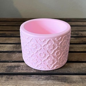 May include: A light pink, cylindrical planter with a floral pattern embossed around the exterior. The planter is made of a solid material and sits on a wooden surface. The interior is smooth and empty.
