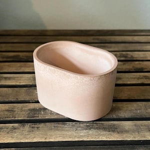 May include: A light pink, oval-shaped planter made of a textured material. The planter has a smooth interior and sits on a wooden surface. The planter is designed for indoor or outdoor use.