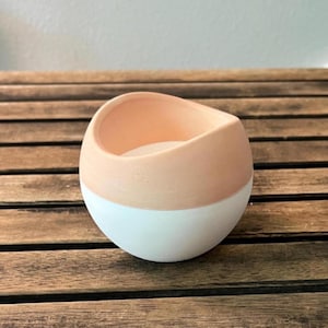 May include: A spherical ceramic bowl featuring a two-tone design with a matte peach-colored upper section and a smooth white lower section. The bowl has an asymmetrical, slanted rim that dips lower on one side, creating an open, rounded vessel shape.