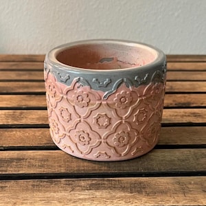 May include: A round, pink and gray ceramic planter with a floral pattern. The planter has a gray rim and a textured surface. The planter is sitting on a wooden surface.