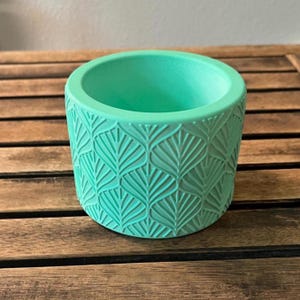 May include: A round, mint-green planter with a textured leaf pattern. The planter is made of a solid material and sits on a wooden surface. The interior is smooth and open, suitable for small plants or decorative items.