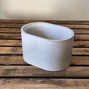 May include: A light gray, oval-shaped concrete planter. The planter has a smooth, textured surface and a rounded top edge. It sits on a wooden surface. The planter is designed for indoor or outdoor use.