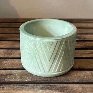 May include: A light green, cylindrical planter with a geometric chevron pattern. The planter is made of a textured material and sits on a wooden surface. The planter has a rounded base and a smooth interior.