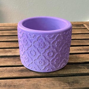 May include: A lavender-colored ceramic planter with a repeating floral pattern. The cylindrical pot has a smooth interior and a textured exterior. The design features stylized flowers and geometric shapes, suitable for indoor or outdoor use.