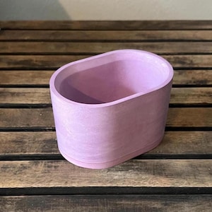 May include: A lavender-colored, oval-shaped planter made of a textured material. The planter has rounded edges and sits on a wooden surface. The interior is empty, suggesting it's ready for plants or decorative items.