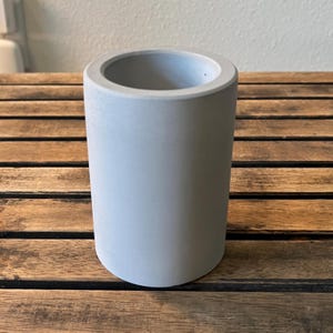 May include: A cylindrical, light gray concrete planter with a smooth surface. The planter has a rounded base and a wide opening at the top. It is sitting on a wooden surface.