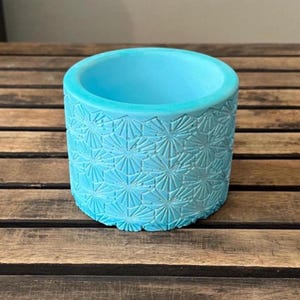 May include: A cylindrical, turquoise-colored planter with a textured, geometric pattern. The planter has a smooth interior and sits on a wooden surface. The design features a repeating starburst pattern around the exterior.