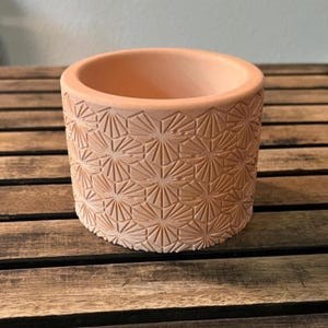 May include: A terracotta-colored cylindrical planter with a geometric, fan-like pattern carved into its exterior. The planter has a wide opening and sits on a wooden surface. Ideal for small plants or succulents.
