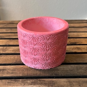 May include: A round, pink planter with a geometric starburst pattern. The planter is made of a textured material and has a smooth interior. The planter is approximately 4 inches in diameter and 3.5 inches tall.