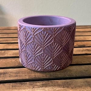 May include: A round, lavender-colored planter with a detailed leaf pattern carved into its exterior. The planter sits on a wooden surface. The interior of the planter is a solid lavender color.