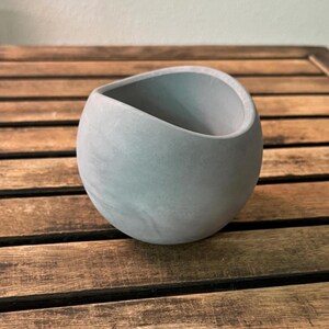 May include: A spherical light gray planter with a smooth, matte finish and a wide, slanted circular opening at the top. The vessel features a solid, rounded base and a clean, continuous exterior surface without any additional patterns or textures.
