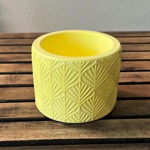 May include: A round, yellow planter with a leaf pattern embossed on the exterior. The interior is also yellow. The planter is sitting on a wooden surface. This decorative planter is suitable for small plants and succulents.