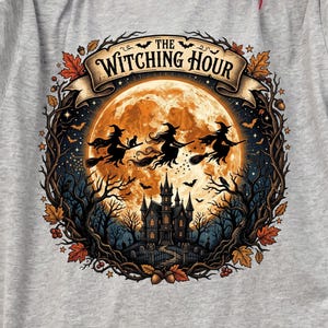 May include: A sport grey t-shirt featuring a graphic of a full moon, a dark castle, and silhouettes of three witches flying on broomsticks. The design is framed by autumn leaves and branches, with the text "THE WITCHING HOUR" in a banner.