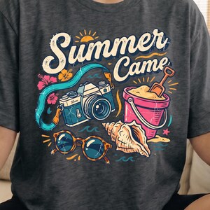 May include: Dark grey heather t-shirt with a colorful graphic that says "Summer Came." The design includes a vintage camera, sunglasses, a seashell, a pink bucket with sand, and a shovel. The shirt is a casual, comfortable style.