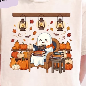 May include: A natural-colored t-shirt featuring a graphic of a white ghost sitting in a wicker chair reading a book titled Spooky Tales. The scene includes stacks of books labeled Potions, Ancient Spells, and Ghostly Lore, surrounded by pumpkins, hanging lanterns, autumn leaves, and bats.