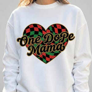 May include: White sweatshirt featuring a heart-shaped design with a red, black, and green checkered pattern. The words "One Dope Mama" are printed in a retro font, outlined in gold and black.