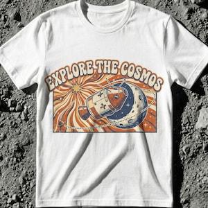 May include: White t-shirt featuring a retro-style graphic with the text "EXPLORE THE COSMOS." The design includes a rocket ship, a planet, and a sunburst background in shades of orange, blue, and brown.