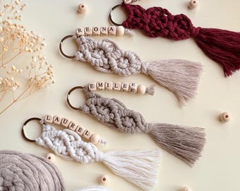 Pode incluir: Chaveiros de macramé feitos à mão em várias cores, incluindo bordô, bege e cinza. Cada chaveiro apresenta um nome escrito com contas de letras de madeira e uma borla. Os chaveiros são presos a um anel de metal.
