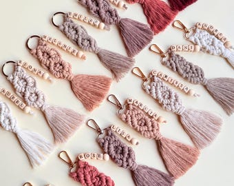 Pode incluir: Uma coleção de chaveiros de macramé feitos à mão em várias cores, incluindo rosa, malva e bordô. Cada chaveiro apresenta um nome escrito com contas de letras de madeira e uma borla. Os chaveiros são presos a fechos dourados.