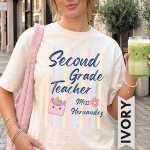 Puede incluir: Una camiseta de manga corta color marfil con un gráfico de rayas verticales en tonos pastel y el texto en azul oscuro Second Grade Teacher Miss Hernandez. El diseño incluye una caja de ceras rosa con cara y una pequeña flor rosa sonriente.