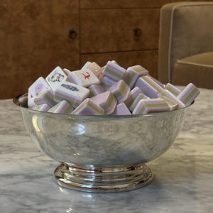 Puede incluir: Un cuenco de metal plateado con pedestal lleno de una colección de fichas de Mahjong. Las fichas presentan un diseño en capas con rayas blancas, verde claro y lavanda, mientras que algunas muestran ilustraciones florales y de jarrones.