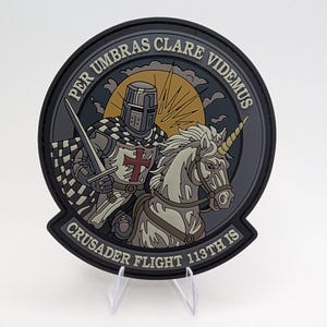 May include: A circular patch with a knight on a unicorn, the text reads "PER UMBRAS CLARE VIDEMUS" and "CRUSADER FLIGHT 113TH IS". The knight is in armour, holding a sword, and the unicorn has a golden horn. The patch has a black border.