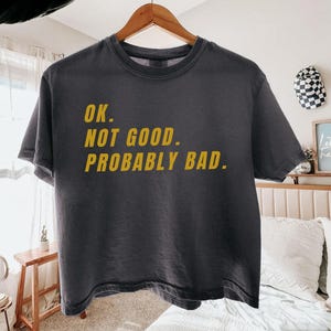 May include: Dark grey t-shirt with the text "OK. NOT GOOD. PROBABLY BAD." in yellow letters. The shirt is hanging on a wooden hanger. The t-shirt is a casual wear item.