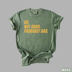 Pu&ograve; includere: Una maglietta verde muschio con la scritta "OK. NOT GOOD. PROBABLY BAD." in giallo. La maglietta ha un girocollo e maniche corte. La maglietta &egrave; realizzata in un materiale morbido e confortevole.