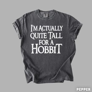 Puede incluir: Camiseta gris oscuro con el texto blanco "I'M ACTUALLY QUITE TALL FOR A HOBBIT". La camiseta tiene cuello redondo y mangas enrolladas. La tela parece suave y ligeramente descolorida.