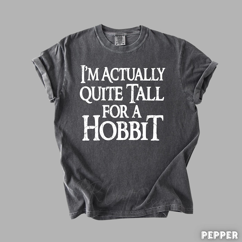 Puede incluir: Camiseta gris oscuro con el texto blanco "I'M ACTUALLY QUITE TALL FOR A HOBBIT". La camiseta tiene cuello redondo y mangas enrolladas. La tela parece suave y ligeramente descolorida.